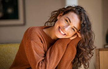 A woman with curly hair wearing a brown sweater sits indoors, smiling and leaning her head on her hands.