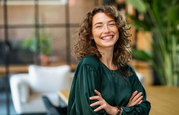 A smiling person with curly hair in a green top stands with arms crossed in a modern room with plants and blurred background.
